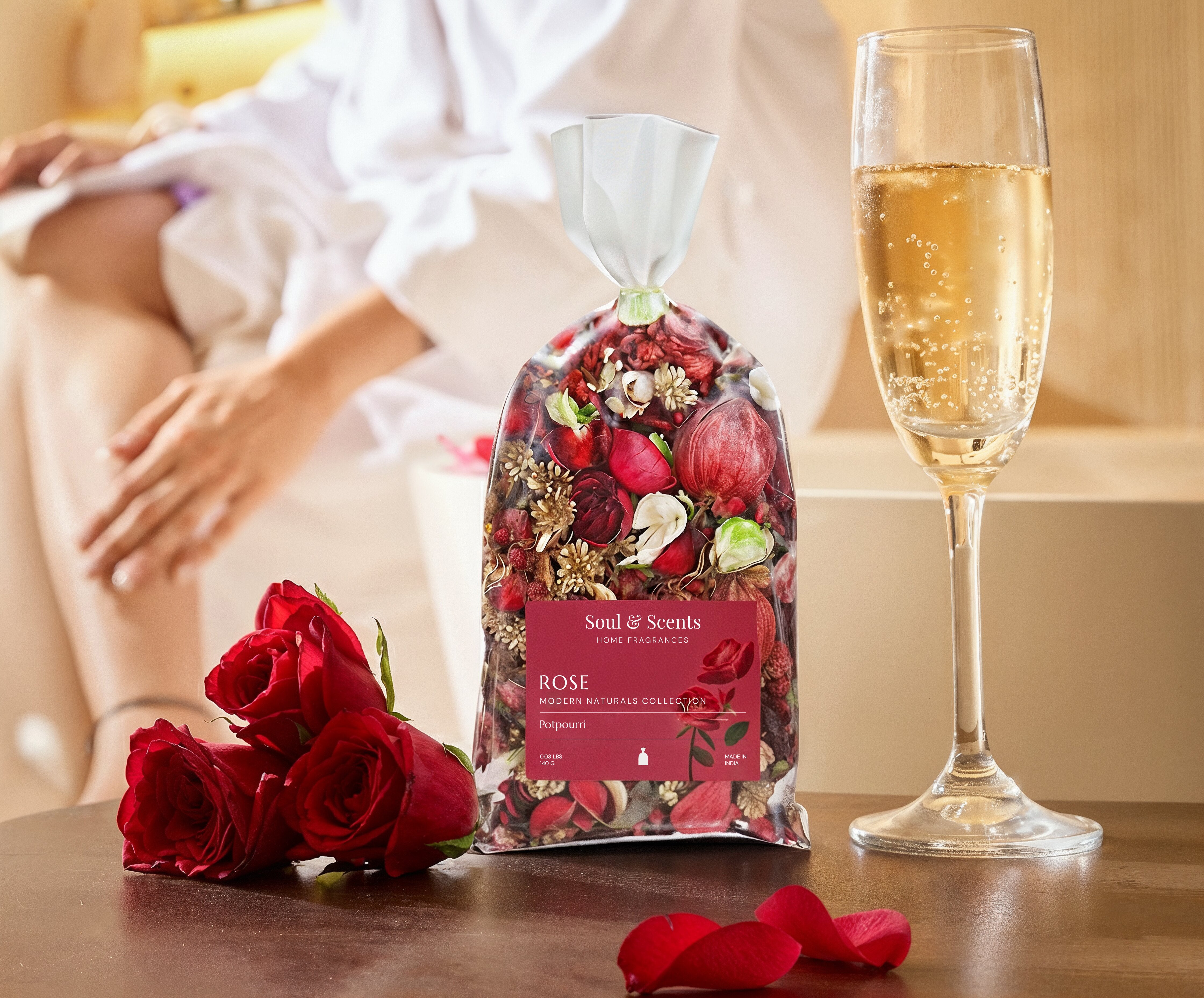 Rose petal bag, red roses, and a glass of champagne on a table with a blurred person in the background.