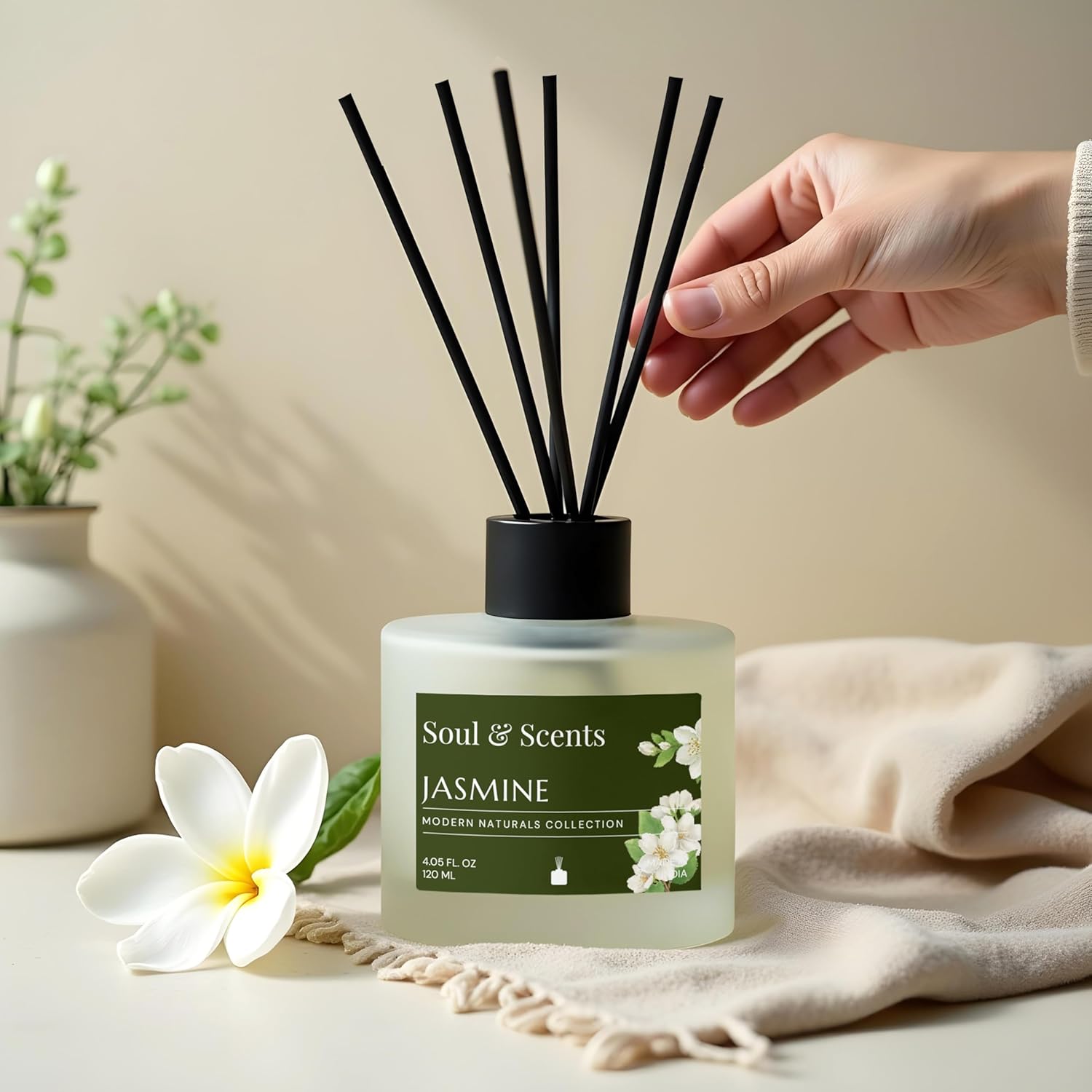 Soul & Scents Jasmine diffuser with a hand holding black sticks, surrounded by flowers and fabric.
