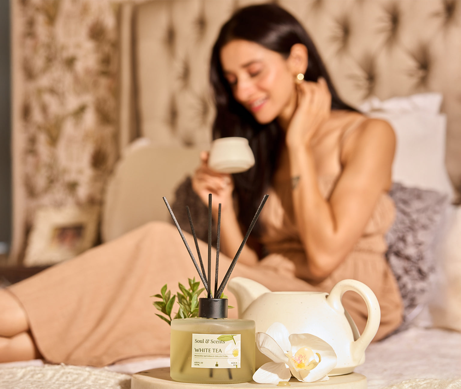 Woman sitting on a bed with a diffuser, teapot, and orchid in a cozy bedroom setting.